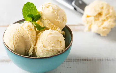 Coppa di cremoso gelato alla vaniglia, guarnita con foglioline di menta fresca. Sullo sfondo, un cucchiaio da gelato pieno.