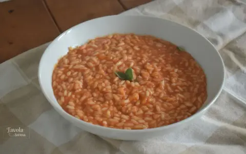 Un cremoso riso al pomodoro veloce, guarnito con una fogliolina verde, servito in una ciotola bianca su una tovaglia a quadri.