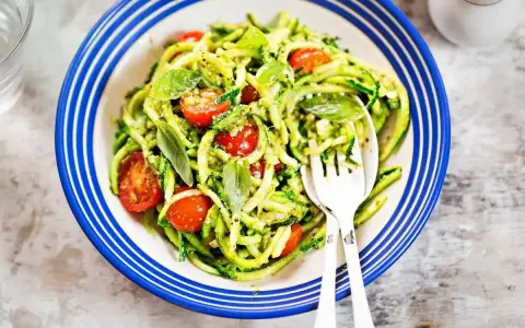 Deliziosi primi piatti con zucchine e pomodorini, conditi con pesto fresco e basilico, serviti in un piatto blu e bianco.