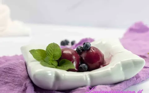 Due palline di gelato ai mirtilli, guarnite con mirtilli freschi e foglie di menta, servite in una ciotola bianca su un tovagliolo viola.