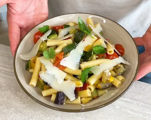 Un piatto di pasta fredda gourmet con verdure miste, pomodorini, basilico e scaglie di parmigiano.