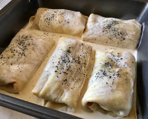 Crespelle farina di ceci ripiene, pronte per essere infornate, immerse in una cremosa besciamella e cosparse di pepe nero.