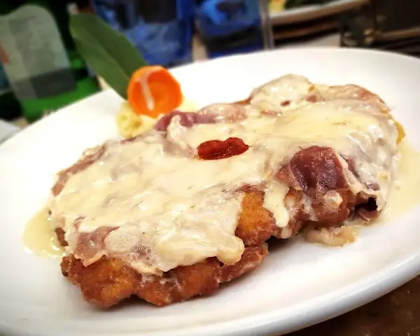 Cotoletta alla bolognese dorata e filante, con prosciutto e formaggio fuso, servita su un piatto bianco con contorno di purè e carota.