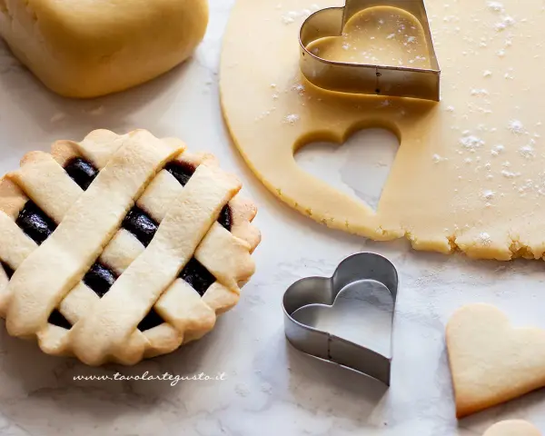 Preparazione di crostatine con pasta frolla classica. Stesa di impasto, formine a cuore e una crostatina già pronta.