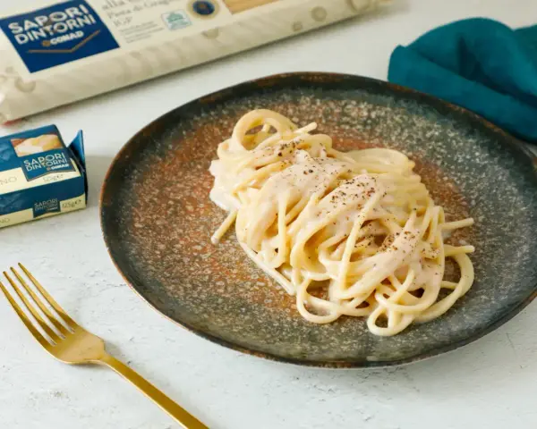 Un piatto di spaghetti cacio e pepe cremosi, guarniti con pepe nero macinato. Accanto, confezioni di pasta e burro, e una forchetta dorata.