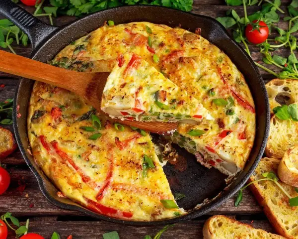 Frittata di peperoni dorata in padella, con fette di pane tostato e pomodorini freschi.