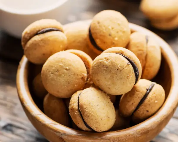 Baci di dama ricetta: deliziosi biscotti alle nocciole con ripieno di cioccolato, serviti in una ciotola di legno.