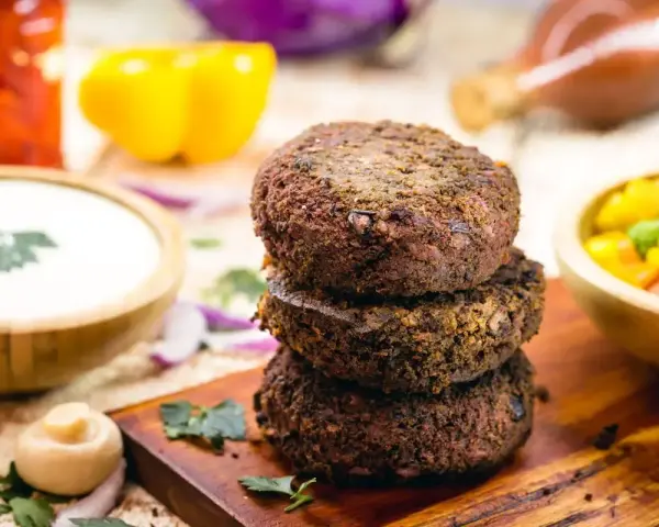 Tre gustosi hamburger di fagioli impilati su un tagliere di legno, con contorni colorati come salsa, peperoni e insalata.