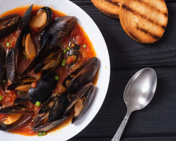 Un invitante piatto di zuppa di cozze in salsa rossa, accompagnato da crostini tostati e un cucchiaio d'argento.