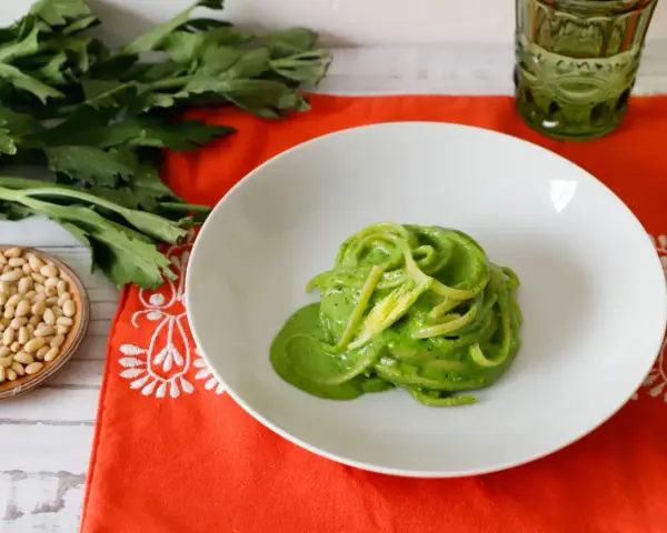 Piatto di spaghetti verdi con salsa cremosa, ispirato a ricette con foglie di sedano. Accanto, pinoli e un bicchiere verde.