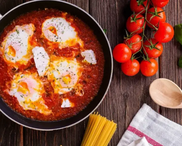 Uova al pomodoro in padella, con pomodorini freschi, basilico e un cucchiaio di legno.