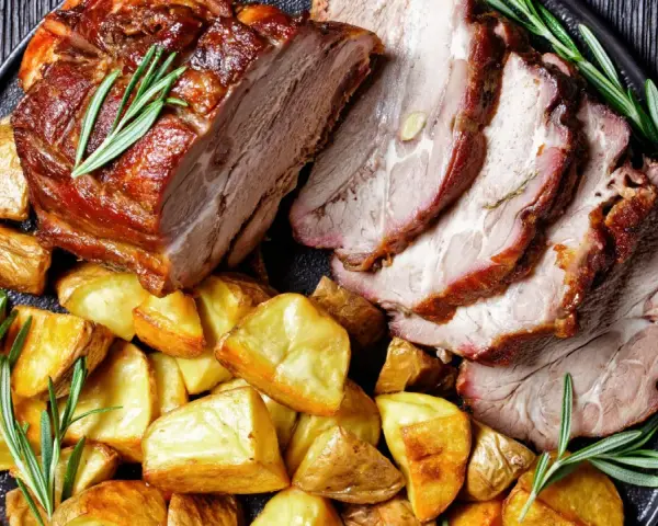 Lombata di maiale arrosto affettata, servita con patate dorate e rametti di rosmarino su un piatto scuro.