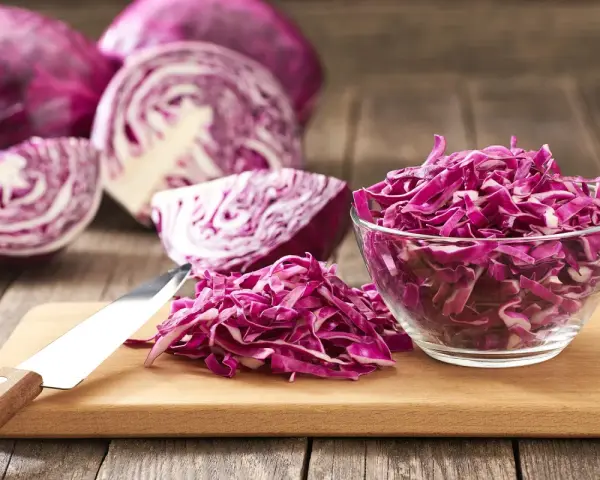 Cavolo cappuccio rosso affettato in una ciotola di vetro e su un tagliere, con cavoli interi e tagliati a spicchi sullo sfondo.