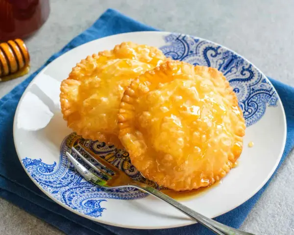 Due dolci sardi fritti, dorati e lucidi di miele, su un piatto blu e bianco con una forchetta.