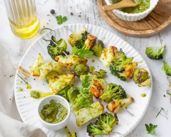 Spiedini di verdure grigliate, con broccoli, cavolfiore e cavolini di Bruxelles, conditi con pesto e serviti su un piatto bianco.