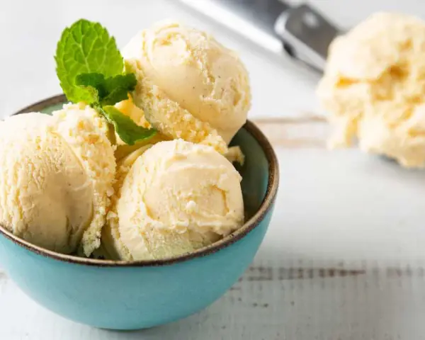 Coppa di cremoso gelato alla vaniglia, guarnita con foglioline di menta fresca. Sullo sfondo, un cucchiaio da gelato pieno.