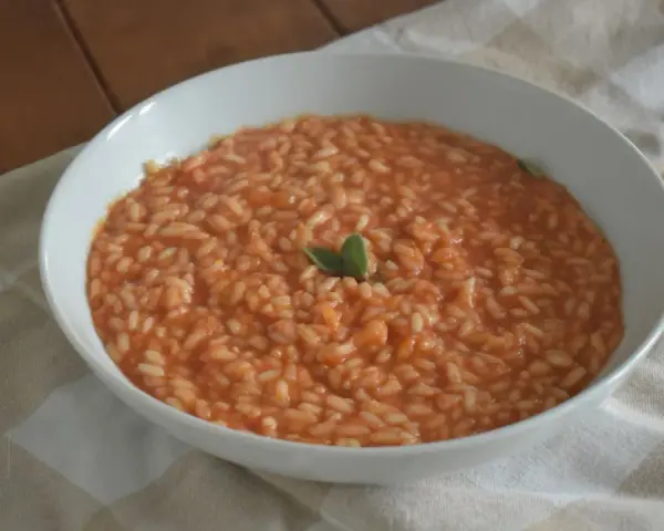 Un cremoso riso al pomodoro veloce, guarnito con una fogliolina verde, servito in una ciotola bianca su una tovaglia a quadri.