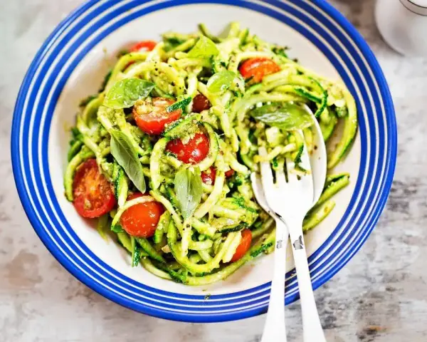 Deliziosi primi piatti con zucchine e pomodorini, conditi con pesto fresco e basilico, serviti in un piatto blu e bianco.