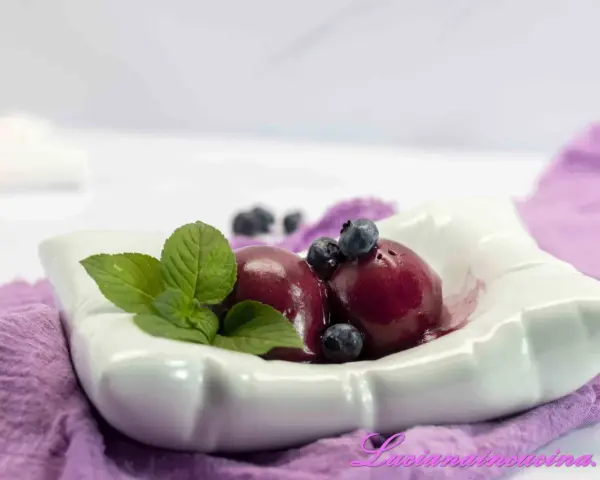 Due palline di gelato ai mirtilli, guarnite con mirtilli freschi e foglie di menta, servite in una ciotola bianca su un tovagliolo viola.