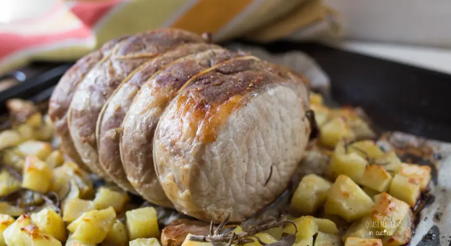 A tied roast pork loin sits on a bed of roasted potatoes and herbs on a baking sheet.