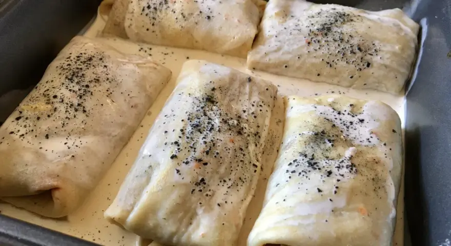 Crespelle farina di ceci ripiene, pronte per essere infornate, immerse in una cremosa besciamella e cosparse di pepe nero.