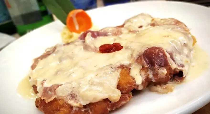Cotoletta alla bolognese dorata e filante, con prosciutto e formaggio fuso, servita su un piatto bianco con contorno di purè e carota.
