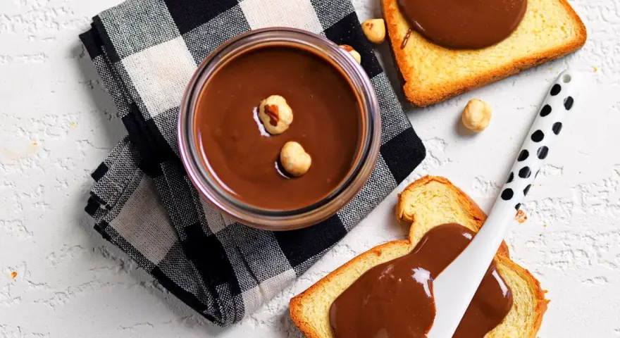 Crema di nocciole fatta in casa in un barattolo, con fette biscottate e nocciole sparse.