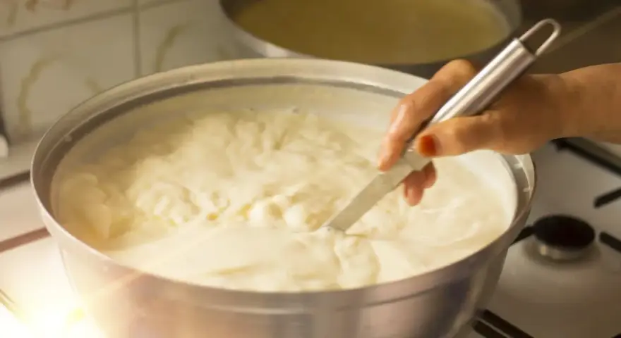 Una mano mescola un cremoso yogurt fatto in casa con fermenti in una ciotola d'acciaio su un fornello.