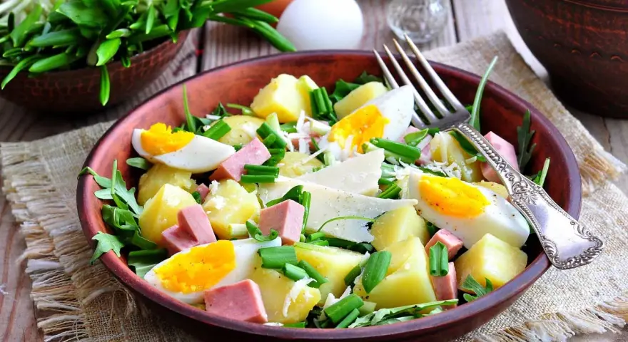 Deliziosa insalata di patate estiva con uova sode, salumi, formaggio e erbe fresche, servita in una ciotola di terracotta.