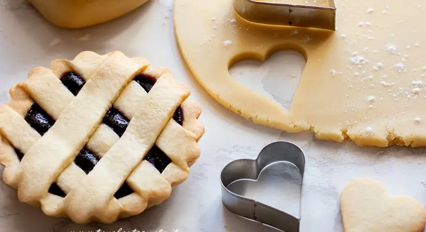 Preparazione di crostatine con pasta frolla classica. Stesa di impasto, formine a cuore e una crostatina già pronta.