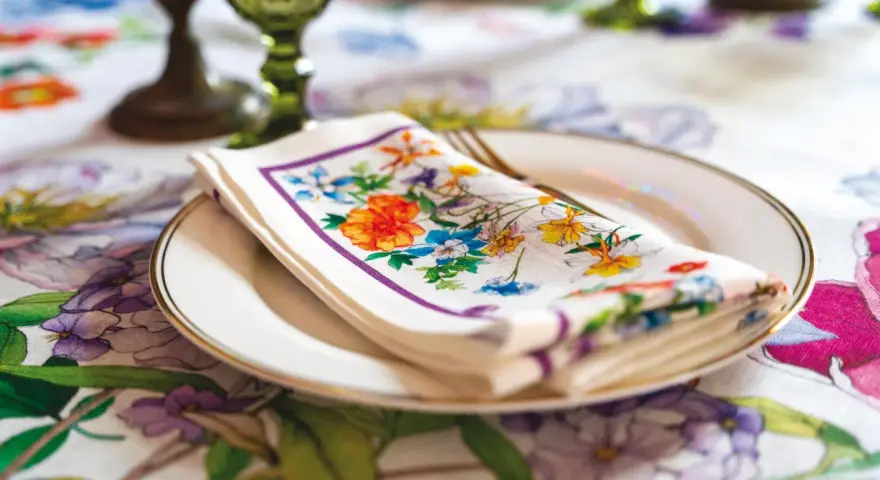 Tovagliolo fiorito piegato su un piatto bianco, con un bordo dorato. La tovaglia è decorata con un motivo floreale vivace. Il tovagliolo va a destra o sinistra?