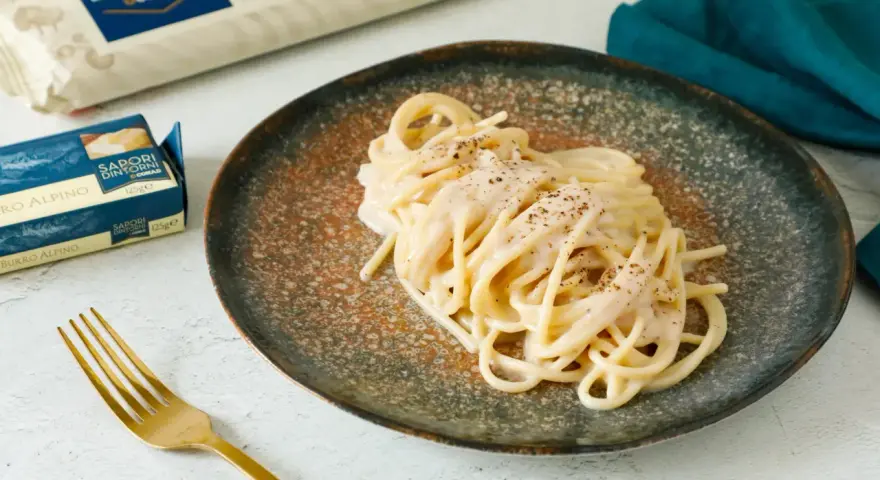 Un piatto di spaghetti cacio e pepe cremosi, guarniti con pepe nero macinato. Accanto, confezioni di pasta e burro, e una forchetta dorata.
