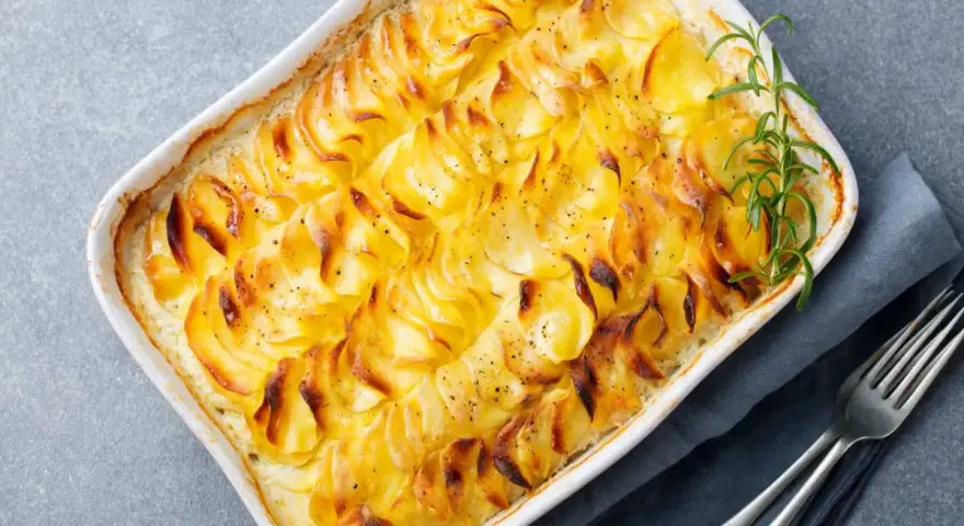Delizioso millefoglie di patate dorate e croccanti, cotto in una teglia bianca, guarnito con rosmarino fresco e pepe nero.