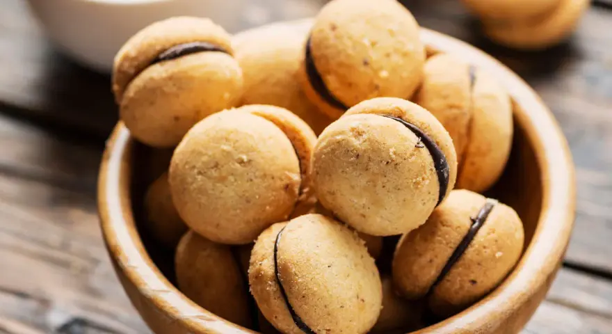 Baci di dama ricetta: deliziosi biscotti alle nocciole con ripieno di cioccolato, serviti in una ciotola di legno.