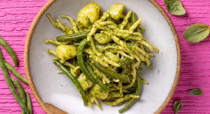 Un piatto invitante di trofie al pesto, con fagiolini e patate, su uno sfondo rosa vibrante.