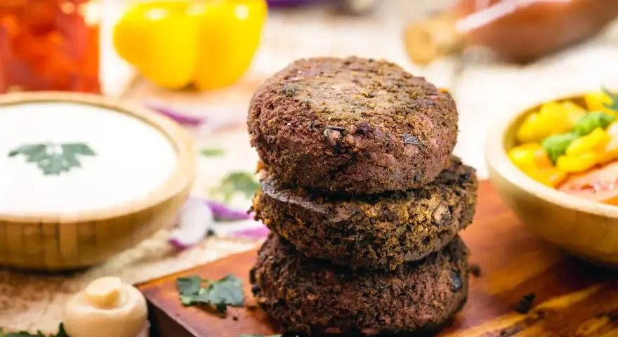 Tre gustosi hamburger di fagioli impilati su un tagliere di legno, con contorni colorati come salsa, peperoni e insalata.