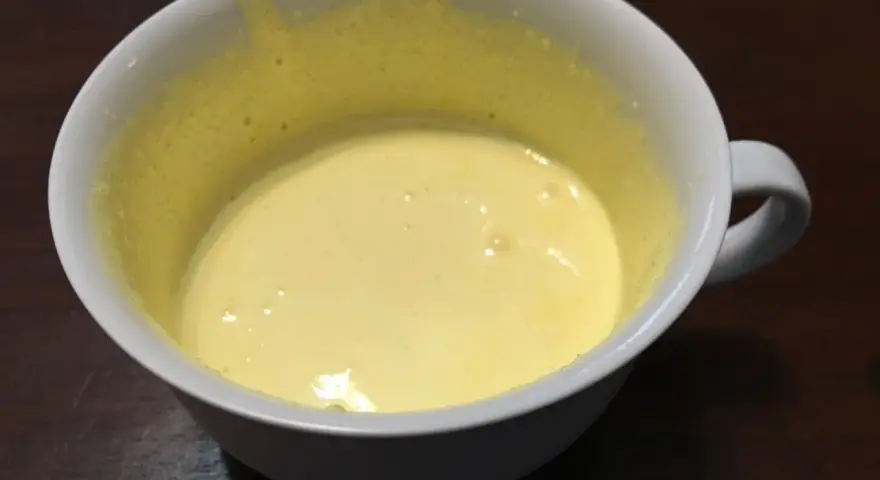 Una tazza bianca contiene un composto giallo pallido, un uovo sbattuto con zucchero, pronto per essere cucinato.