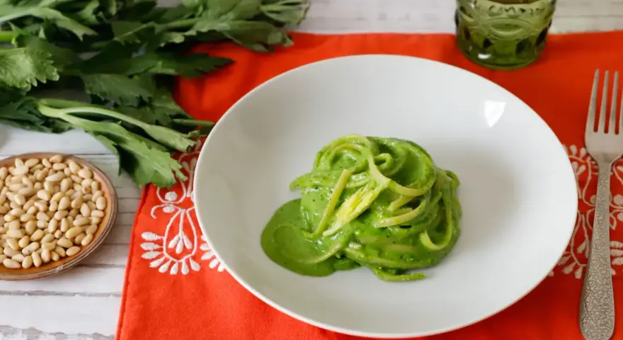 Piatto di spaghetti verdi con salsa cremosa, ispirato a ricette con foglie di sedano. Accanto, pinoli e un bicchiere verde.