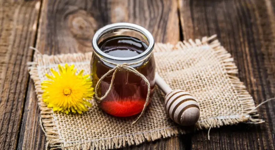 Un vasetto di miele ambrato, un fiore giallo e un cucchiaio di legno su tela grezza. La differenza tra miele e melata è un mistero da svelare.