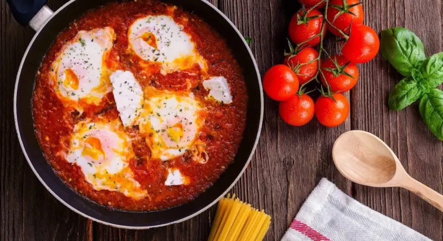 Uova al pomodoro in padella, con pomodorini freschi, basilico e un cucchiaio di legno.