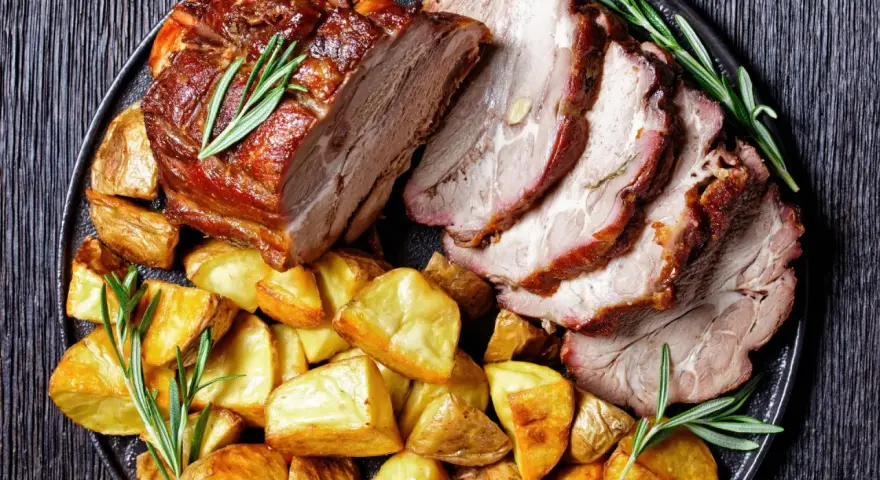 Lombata di maiale arrosto affettata, servita con patate dorate e rametti di rosmarino su un piatto scuro.
