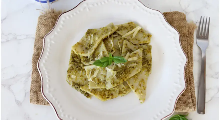 Testaroli al pesto, un piatto tradizionale ligure, servito su un piatto bianco con formaggio grattugiato e basilico fresco.