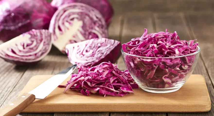 Cavolo cappuccio rosso affettato in una ciotola di vetro e su un tagliere, con cavoli interi e tagliati a spicchi sullo sfondo.