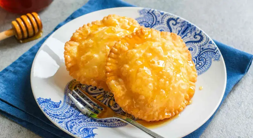 Due dolci sardi fritti, dorati e lucidi di miele, su un piatto blu e bianco con una forchetta.