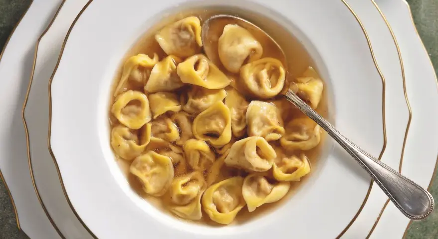 Un primo piatto fumante: tortellini in brodo, un classico della tradizione culinaria italiana, serviti in una ciotola bianca con bordo dorato.