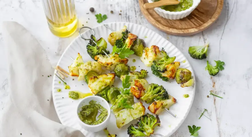 Spiedini di verdure grigliate, con broccoli, cavolfiore e cavolini di Bruxelles, conditi con pesto e serviti su un piatto bianco.