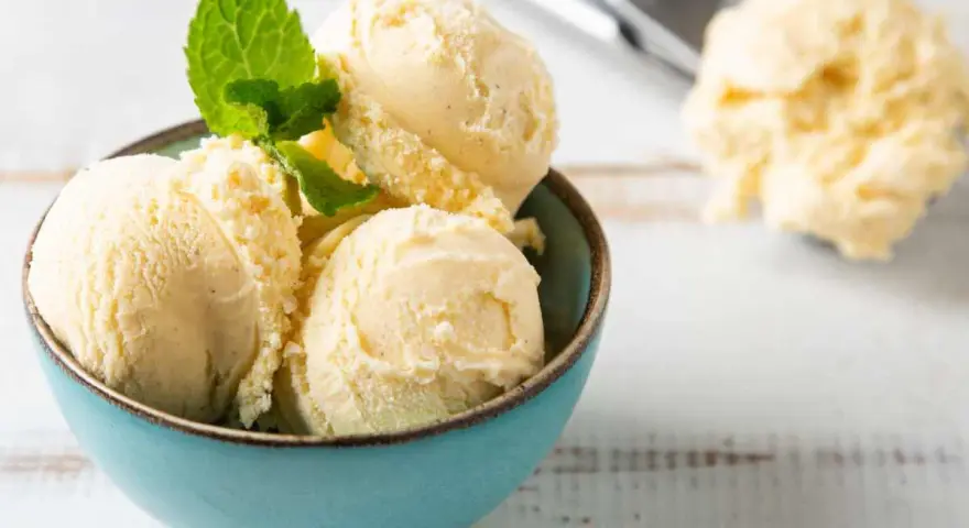 Coppa di cremoso gelato alla vaniglia, guarnita con foglioline di menta fresca. Sullo sfondo, un cucchiaio da gelato pieno.