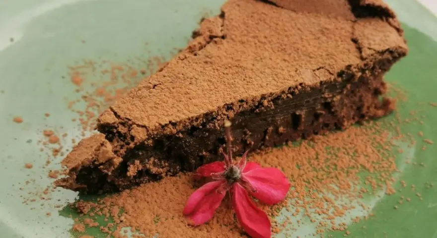 Una fetta di torta tenerina vegana, cosparsa di cacao, con un fiore rosso su un piatto verde.
