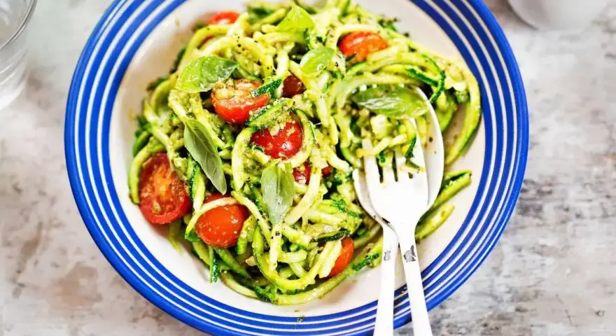 Deliziosi primi piatti con zucchine e pomodorini, conditi con pesto fresco e basilico, serviti in un piatto blu e bianco.