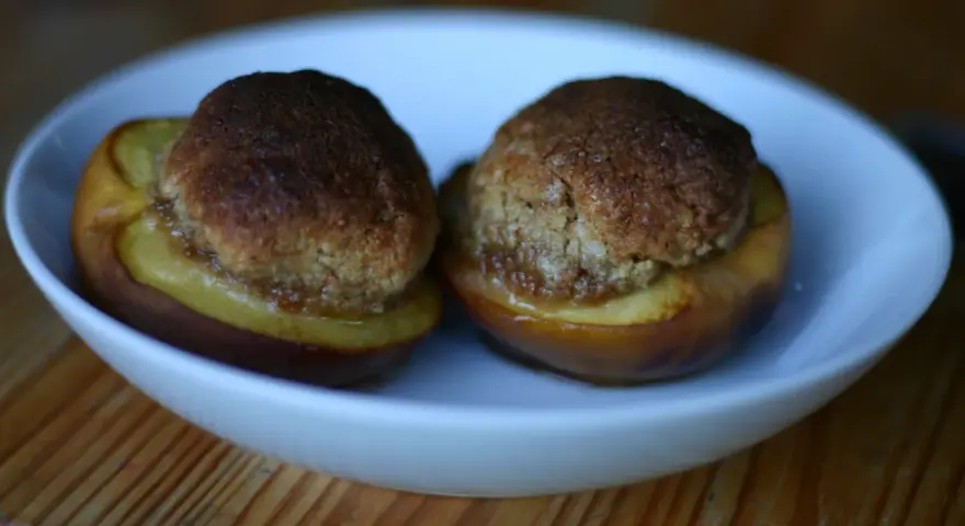 Due pesche ripiene senza amaretti, cotte al forno, servite in una ciotola bianca su un tavolo di legno.